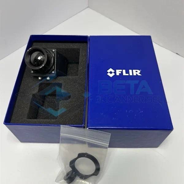 Top-down view of the FLIR A65 thermal camera kit in its original box packaging, placed on a white surface. On the left, the compact black FLIR A65 camera rests inside its custom foam-padded blue box, clearly showcasing its front lens marked "25 mm". Next to it is the original royal blue FLIR branded box cover. In the foreground, a small clear plastic bag contains a black lens adjustment tool / focus ring key accessory.