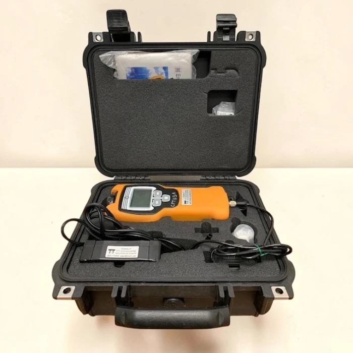An Environics ChemPro100i portable chemical detector, with an orange body and black screen, rests securely inside its custom-foamed black protective carrying case. The lid is open, revealing the device, power cords, and a battery pack.