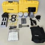 A complete kit for the Trimble Dual R12i GNSS system laid out on a light surface, featuring two yellow hard cases, dual receiver units, a data controller, charging docks, batteries, cables, antennas, and manuals.