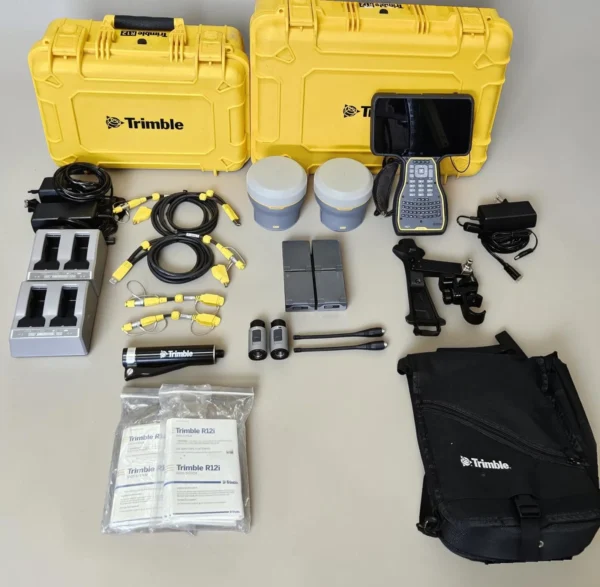 A complete kit for the Trimble Dual R12i GNSS system laid out on a light surface, featuring two yellow hard cases, dual receiver units, a data controller, charging docks, batteries, cables, antennas, and manuals.