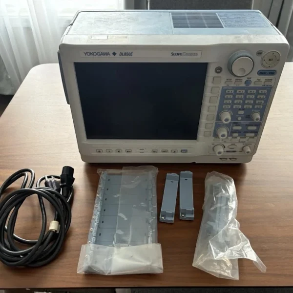 Front view of a Yokogawa DL850E ScopeCorder resting on a wooden table, showing its large screen and control panel, accompanied by a power cord and packaged blank module covers.